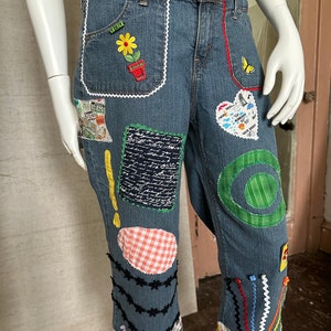 May include: Blue denim jeans with various colorful patches and embroidery, including a flower, a butterfly, a heart, and a circle. The jeans have a white lace trim and black stitching.