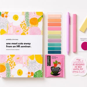 May include: A book titled probably a love story one meet cute away from an HR seminar with a pink floral cover, a set of colorful adhesive page flags, a light purple pen, a pink marker, a yellow eraser, a tea bag, and a sticker with text.