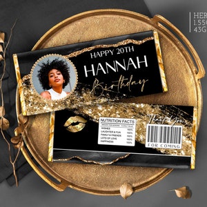 May include: A black and gold chocolate bar wrapper with a photo of a person, the text "Happy 20th Hannah Birthday", and a nutrition facts label. The wrapper is decorated with gold glitter and a gold lip design.