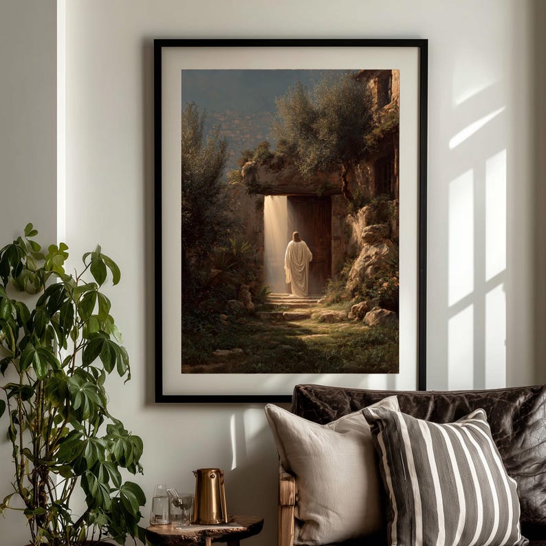 May include: Framed art print depicting a figure in white robes exiting a stone doorway, bathed in sunlight. The artwork features a serene landscape with lush greenery and a dark, ornate frame. The overall theme is religious.