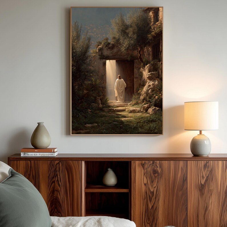 May include: A framed art print depicts a figure in a white robe emerging from a stone doorway, bathed in light. The artwork, with a warm color palette, is set in a natural, outdoor environment with lush greenery and stone steps. The print is displayed above a wooden cabinet.