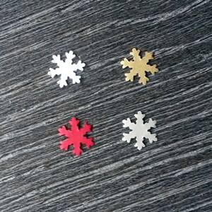 May include: Four snowflake-shaped ornaments in white, gold, red, and silver. The ornaments are arranged on a dark gray, textured surface. These decorative snowflakes are ideal for holiday crafts and winter-themed decor.