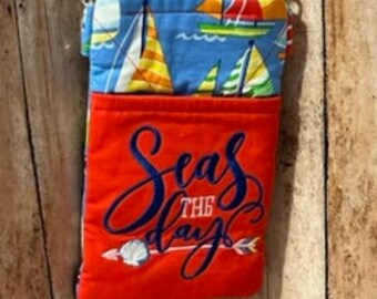 May include: Red and blue fabric crossbody bag with a sailboat print. The front pocket has the text "Seas the day" embroidered in white and blue.