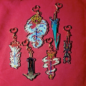 May include: Five colorful keychains featuring different designs. Each keychain has a gold-colored metal ring and a clear acrylic charm. The charms feature a variety of designs, including a sword, a staff, and a flower.