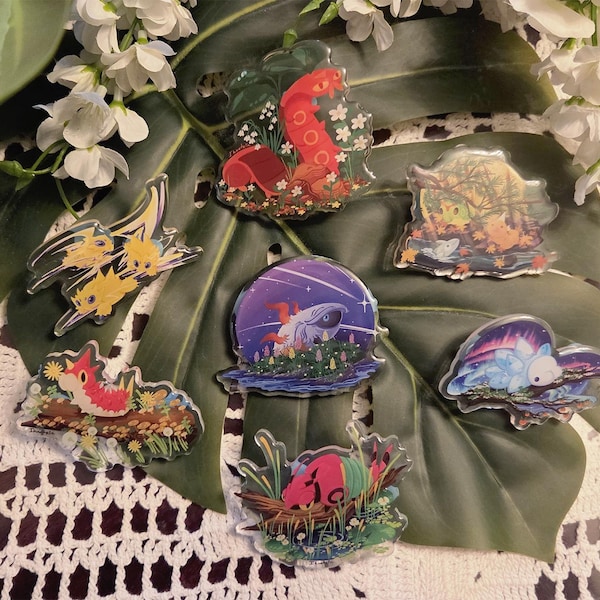 May include: A collection of seven clear acrylic pins featuring colorful illustrated creatures in various natural settings, including a red snake, a yellow flying creature, a blue whale, and a pink animal, all with detailed artistic backgrounds.