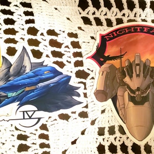 Könnte beinhalten: Zwei Aufkleber mit Illustrationen von futuristischen Robotern. Der erste Aufkleber ist blau und grau mit einem wolfartigen Kopf und dem Text "IV". Der zweite Aufkleber ist grau und zeigt einen Roboter vor einem roten und orangefarbenen Sonnenuntergang mit dem Text "NIGHTFALL".