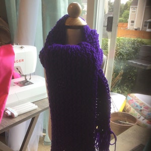 May include: A purple knitted scarf with long fringe on a mannequin. The scarf is made of a chunky yarn and has a loose, open weave.