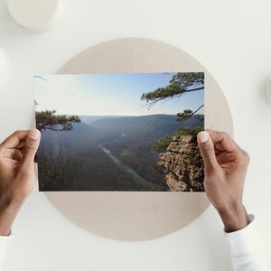 May include: A photograph of a scenic view of a valley with a river winding through it. The photo is held in the hands of a person.