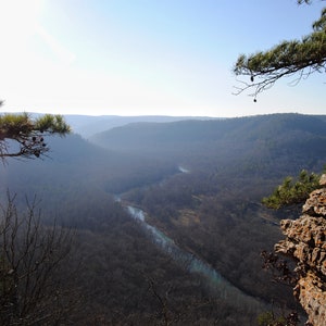 May include: A panoramic view from a clifftop overlooking a valley with a winding river running through it. The trees are bare and the sky is a clear blue.