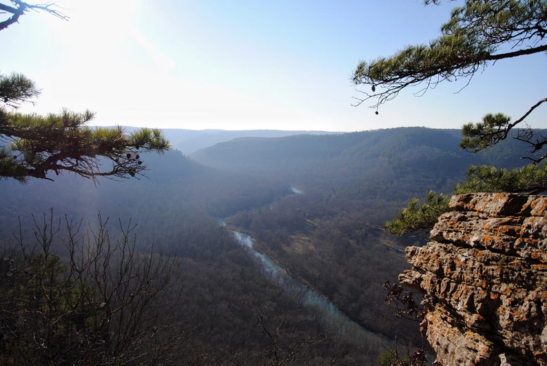 May include: A panoramic view from a clifftop overlooking a valley with a winding river running through it. The trees are bare and the sky is a clear blue.