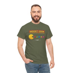 Puede incluir: Camiseta verde oliva con un diseño de Pac-Man y el texto "INSERT COIN" en un recuadro rojo y dorado. El diseño incluye Pac-Man, fantasmas y el texto "+1UP....".
