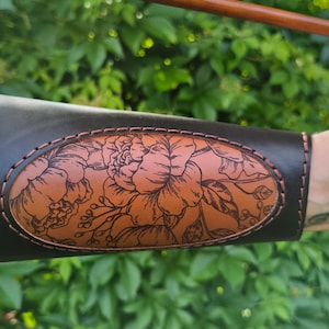 May include: Black leather archery arm guard with a floral design in a lighter brown color. The arm guard features an oval-shaped design with a detailed flower and leaf pattern, stitched with a contrasting thread.