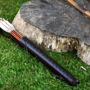 May include: A leather quiver with a brown strap and a set of arrows with white fletching. The quiver is lying on a bed of green grass next to a tree stump.