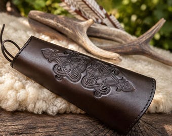 May include: A dark brown leather arm guard featuring an embossed intricate knotwork pattern and thin leather lacing at the top. The surface of the leather is smooth with a polished finish, showcasing detailed symmetrical carvings along the center.