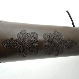 May include: Brown leather arm guard with a Celtic knot design. The guard has a strap with a buckle to adjust the fit.