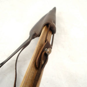 May include: A close-up of a brown leather bow quiver with a wooden arrow shaft and a brass buckle. The quiver is attached to the arrow shaft with leather straps.