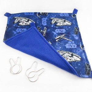 May include: A blue and black Star Wars themed pet hammock with two silver clips. The hammock is made of soft fleece and has a printed design of spaceships and the words "Star Wars".