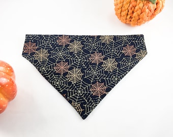 Könnte beinhalten: Dreieckiges schwarzes Haustier-Bandana mit einem sich wiederholenden Muster aus goldenen und orangefarbenen Spinnweben. Das Bandana ist dreieckig gefaltet. Der Hintergrund enthält zwei dekorative Kürbisse.