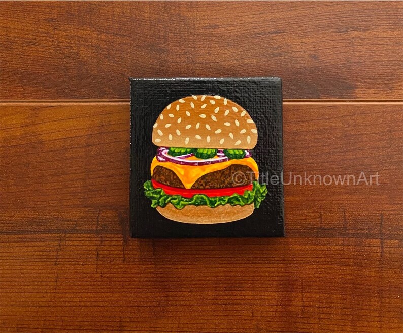 Tiny 2 inch by 2 inch painting. Acrylic on canvas with shiny varnish on top. Features a hamburger with a black background