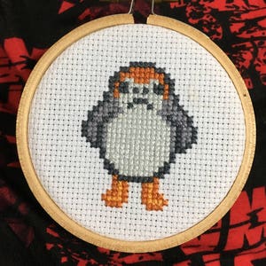Peut inclure: Un cerceau de broderie blanc avec un motif de point de croix représentant un petit personnage orange, gris et blanc avec des pieds orange. Le personnage est un personnage fictif populaire de la franchise Star Wars.