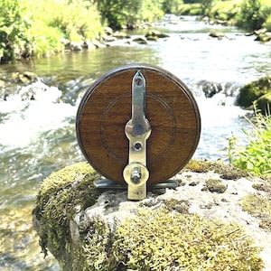 May include: A classic wooden fishing reel with a silver metal mechanism. The reel has a circular, dark wood body and is set against a natural backdrop of a mossy rock and a flowing stream, evoking a sense of traditional angling.