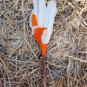 May include: A handmade feather fan featuring white and orange feathers. The feathers are secured to a wooden handle with orange thread. The fan is presented on a bed of dry straw.