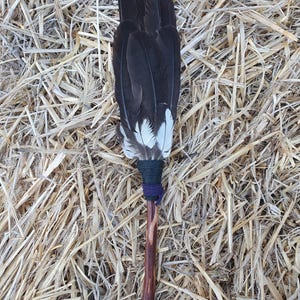 May include: A handcrafted feather fan with a dark wooden handle. The fan features a bundle of black feathers with white tips, bound with dark green and purple twine. The fan rests on a bed of dry straw.