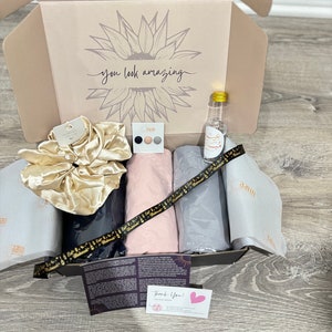 May include: A gift box filled with items including a hair scrunchie, a small bottle of liquid, and two pieces of fabric. The box is lined with white tissue paper with a sunflower design and the text "you look amazing". A thank you card with a pink heart is visible in the bottom right corner.