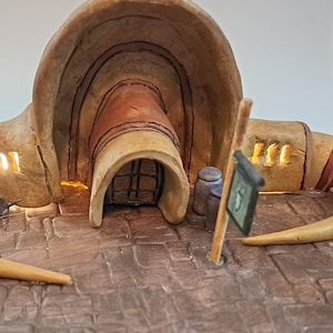 May include: A miniature clay model of a building with a curved roof and two large horns. The building has a door and windows, and the horns are painted a light brown colour.