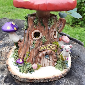 Könnte beinhalten: Ein fantasievolles Baumstumpfhaus mit einem roten Pilzdach, lila und rosa Pilzen und einem Schild mit der Aufschrift "MOG". Eine kleine weiße Maus mit Flügeln sitzt auf einem Pilz in der Nähe des Hauses.