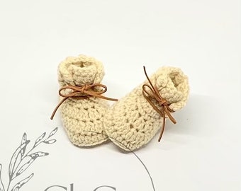 May include: A pair of cream-colored crocheted baby booties with brown leather ties. The ties are tied in a bow at the top of each bootie.