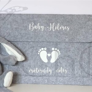 May include: A grey felt folder with a white inscription that reads "Baby Holmes" and a pair of white footprints with a small heart in between. The text "maternity notes" is also written in white below the footprints.