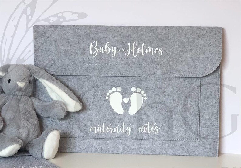 May include: A grey felt folder with a white inscription that reads "Baby Holmes" and a pair of white footprints with a small heart in between. The text "maternity notes" is also written in white below the footprints.