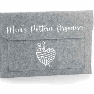 以下が含まれることがあります： グレーのフェルト製パターンオーガナイザーで、"Mom's Pattern Organizer"というテキストと、糸と編み針がついたハートの白いアウトラインがあります。
