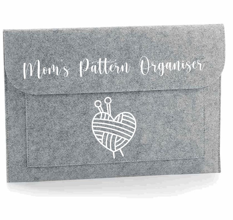 K&ouml;nnte beinhalten: Graue Filz-Aufbewahrungsh&uuml;lle mit dem Text "Mom's Pattern Organiser" und einer wei&szlig;en Umrandung eines Herzens mit Garn und Stricknadeln.