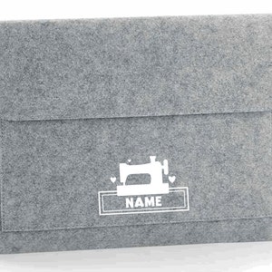 K&ouml;nnte beinhalten: Graue Filz-Laptoptasche mit einem wei&szlig;en N&auml;hmaschinen-Design und dem Wort "NAME" in einem wei&szlig;en Rechteck.