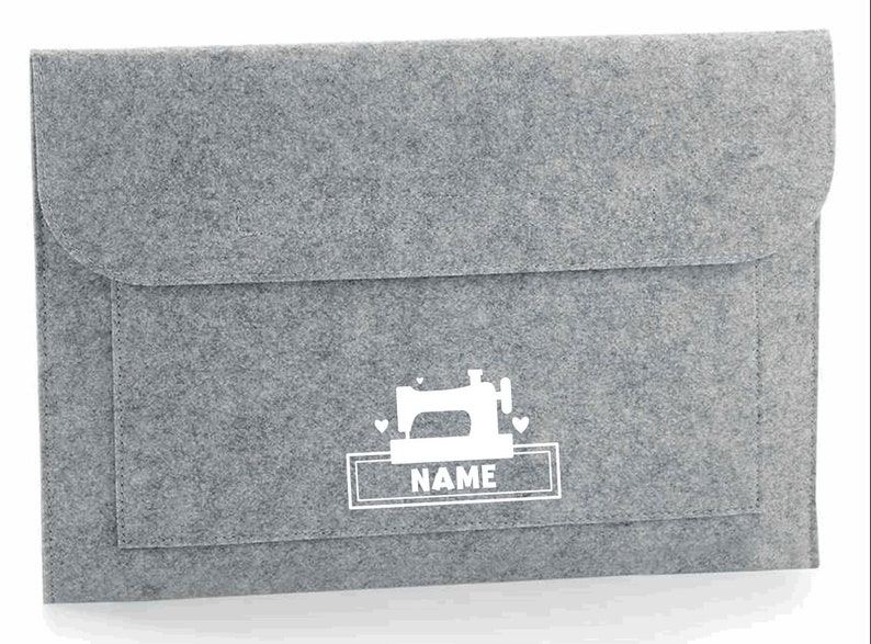K&ouml;nnte beinhalten: Graue Filz-Laptoptasche mit einem wei&szlig;en N&auml;hmaschinen-Design und dem Wort "NAME" in einem wei&szlig;en Rechteck.