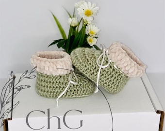 May include: A pair of handmade baby booties in a soft green crochet with a plush, light pink cuff. White laces thread through the front. The booties rest on a white box with the text "ChG CrochetHookersGifts" and are decorated with daisies.