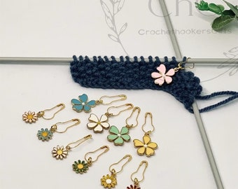 May include: A set of ten colorful flower-shaped stitch markers for knitting or crocheting. The markers are made of metal with a gold finish and have different colored enamel flowers. The markers are shown on a white background with a piece of blue knitted fabric and knitting needles.