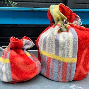 Peut inclure: Deux sacs en feutre, un grand et un petit, sont présentés côte à côte. Le grand sac est principalement rouge, avec une section rayée jaune, grise et bleue. Le petit sac a un design similaire. Les deux sacs sont noués avec de la ficelle.