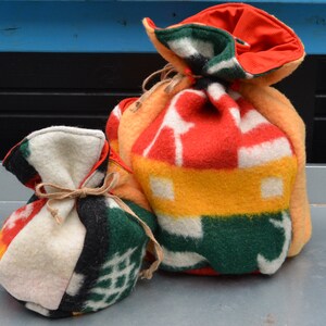 Op de afbeelding: Twee handgemaakte trekkoordtassen. De kleinere tas is gemaakt van een patchwork van kleurrijke stoffen, vastgebonden met touw. De grotere tas is gemaakt van een rood, geel en wit patroonstof, met een oranje voering en trekkoordsluiting.