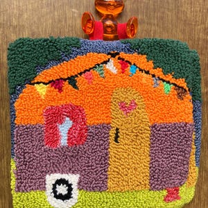 Puede incluir: Un colorido tapiz de pared hecho a mano que representa una casa caprichosa. El diseño presenta un techo naranja, una puerta amarilla y un césped verde. La casa está adornada con una guirnalda de banderas coloridas y un detalle en forma de corazón. Fijado a una ventosa transparente.