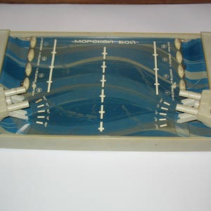 May include: A vintage blue and white plastic game board with the text "Морской Бой" printed on the top. The game board has two white plastic cannons on either side.