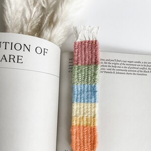 May include: A handwoven bookmark with a striped design in shades of pink, green, blue, yellow, and orange. The bookmark has white fringe at the top and bottom. It is placed on the pages of an open book, adding a touch of colour.