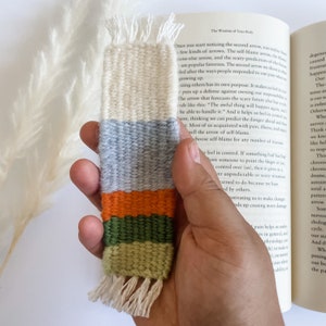 May include: A hand holds a woven bookmark with a white, grey, orange, green, and tan colour scheme. The bookmark has a fringe at the bottom.