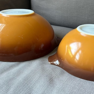 May include: Two vintage Pyrex mixing bowls in a warm amber color with a brown base. The bowls have a rounded shape with a white rim and small handles. These bowls are a classic kitchenware item.