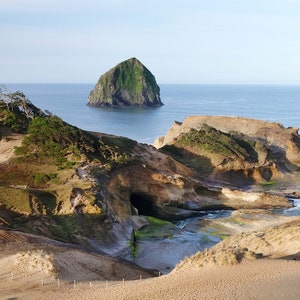 Könnte beinhalten: Panoramablick auf eine felsige Küste mit einer kleinen, grünen Insel in der Ferne. Im Vordergrund befindet sich ein Sandstrand mit einer kleinen Höhle und einem felsigen Ausläufer. Der Himmel ist blau und das Wasser ist tiefblau.
