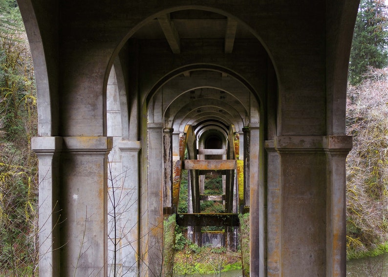K&ouml;nnte beinhalten: Eine Ansicht einer Betonbr&uuml;cke mit mehreren B&ouml;gen, die einen tunnelartigen Effekt erzeugen. Die B&ouml;gen sind in einer Reihe angeordnet und f&uuml;hren zu einer dunklen &Ouml;ffnung am Ende. Die Br&uuml;cke ist aus rauer Beton und hat ein verwittertes Aussehen.
