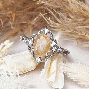 May include: A silver ring with a large, oval-shaped, light brown gemstone surrounded by smaller clear gemstones. The ring has a delicate, ornate design.