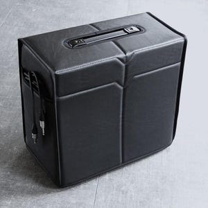 May include: A black, leather-effect box with a handle and side pocket. The box features a geometric design with stitched seams. Cables extend from the side. Likely a speaker or amplifier.